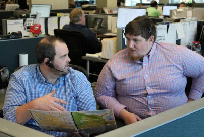 Two men sitting at a desk wearing headsets. One holds a brochure and gestures to the other.