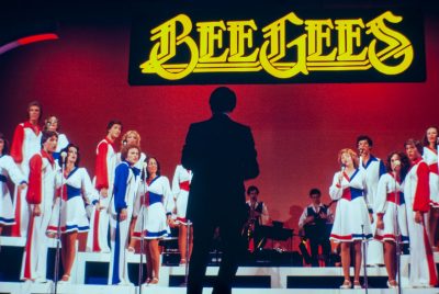 A man stands in front of a group of performers onstage who are dressed in red, white, and blue costumes - women in dresses and men in slacks, shirts, and vests. Some are singing into microphones, while others are playing instruments. A huge lighted sign above them reads "Bee Gees"