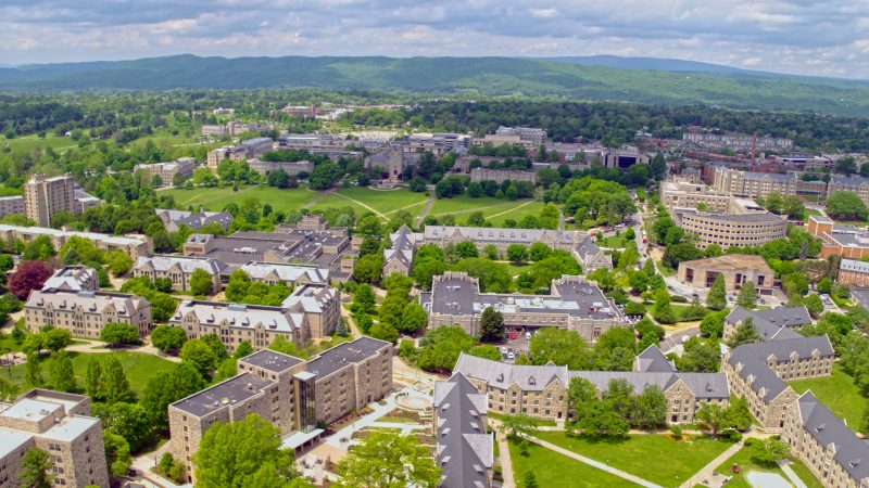 From an aerial view, gray Hokie Stone buildings are tucked between greenery in a mountain valley on a clear, sunny day.