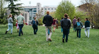 A group of people walk away from the camera while following along for a talking walk about pollinators on campus. Lane Stadium is in the background.